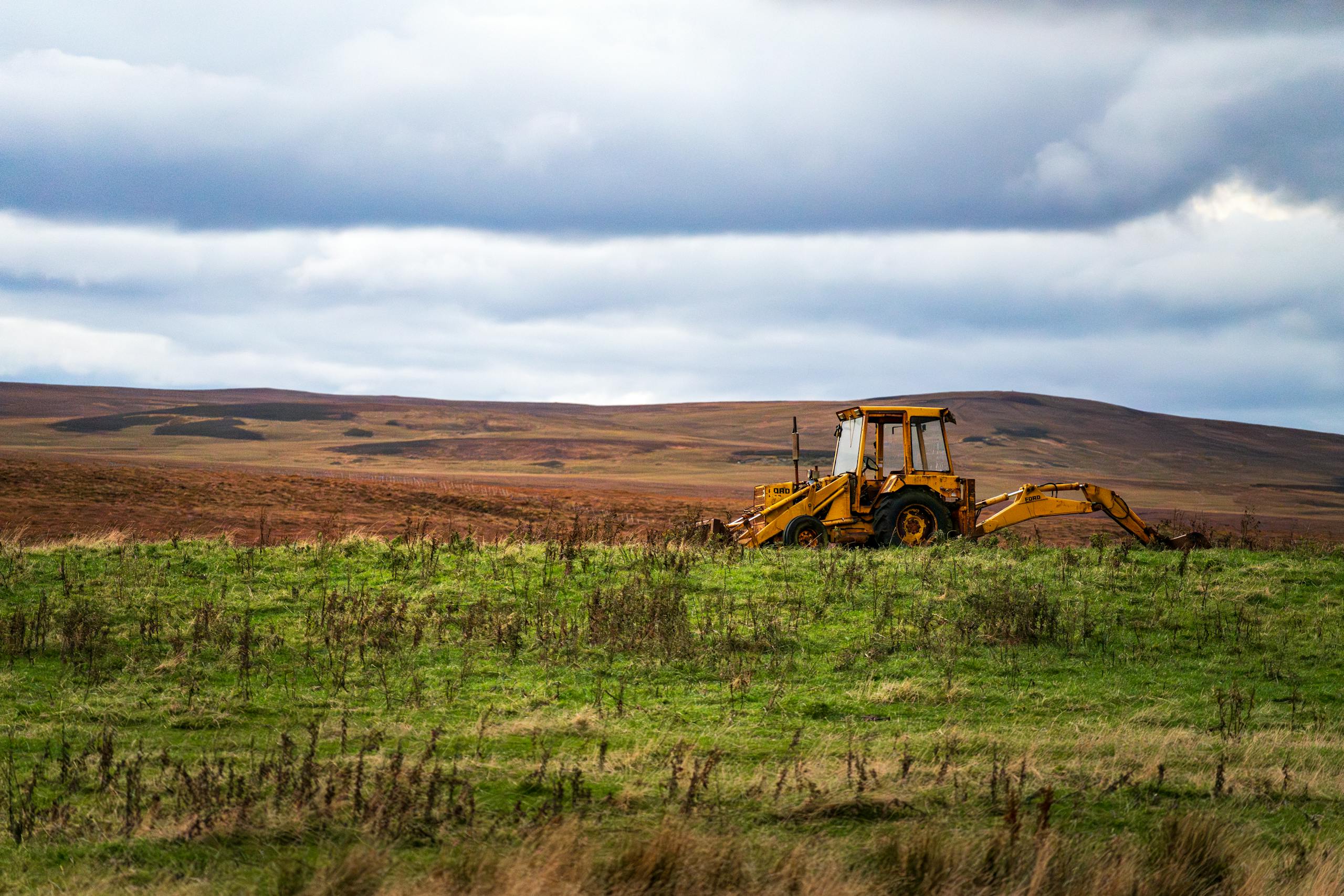 A solitary yellow backhoe on a wide green field against a backdrop of hills and a cloudy sky.