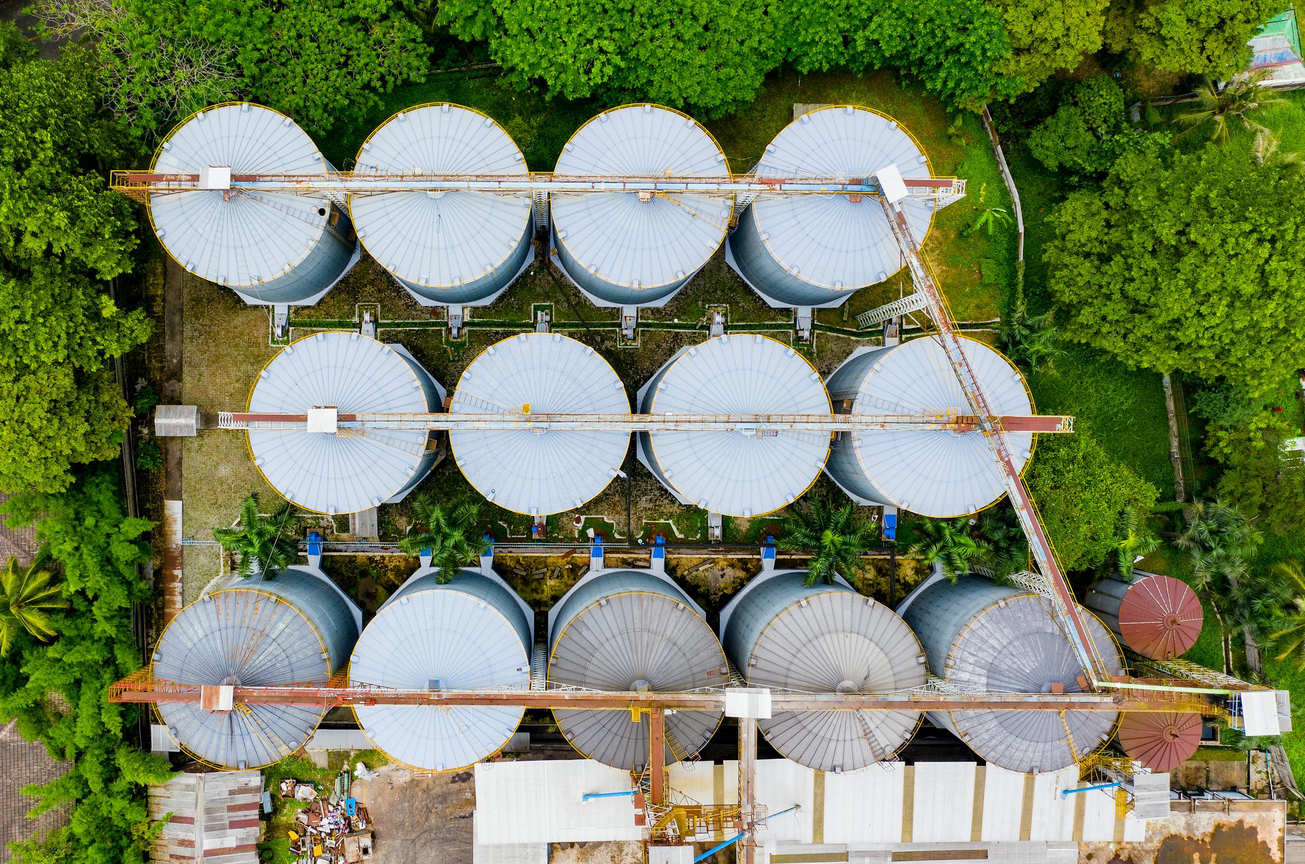 Aerial shot of metal storage tanks surrounded by green foliage in an industrial plant.